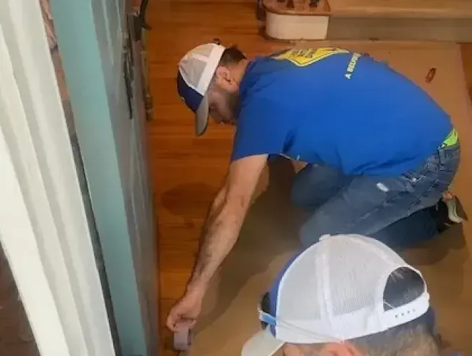 Two workers kneeling on a wood floor to check water damage along the baseboard